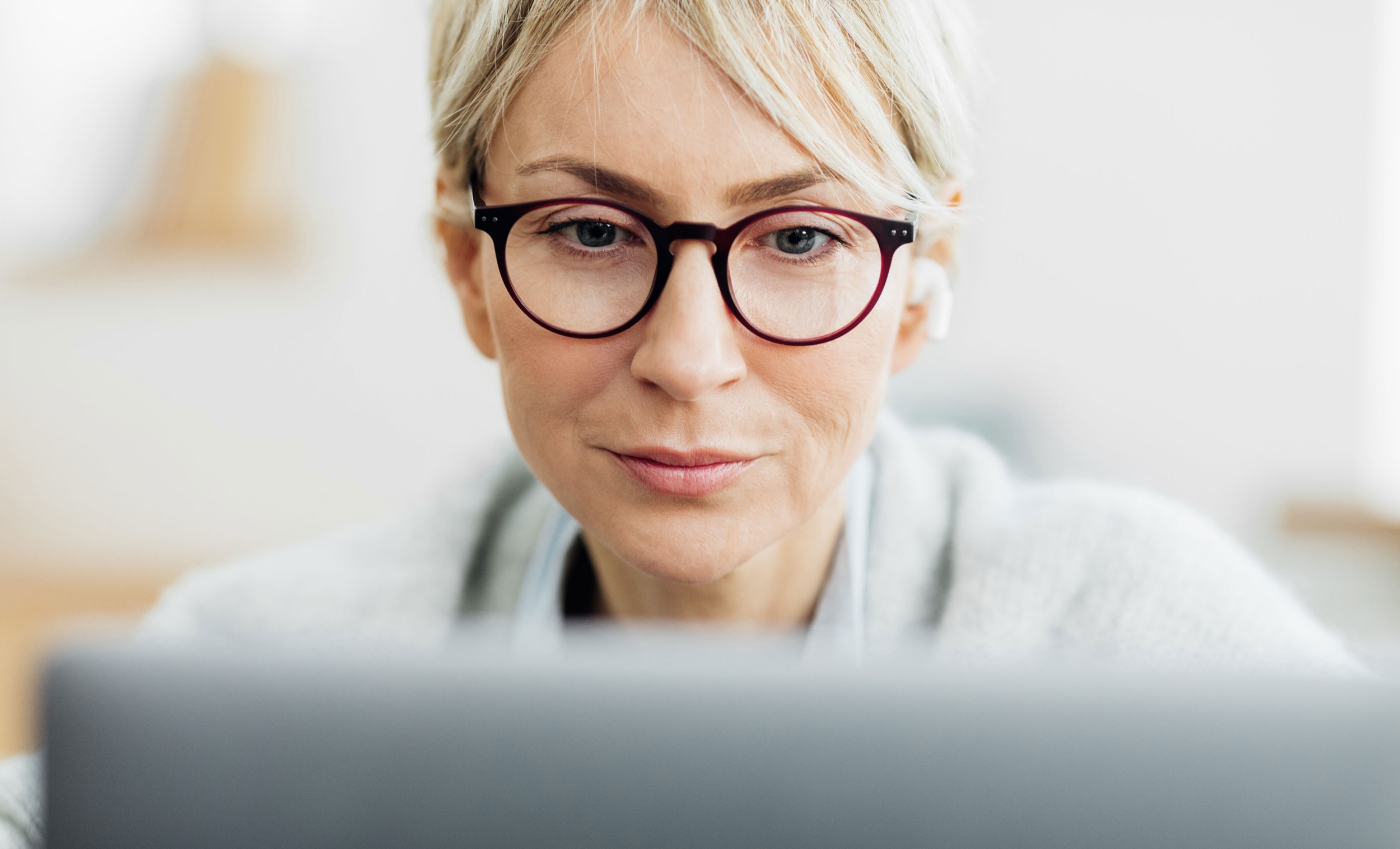 A middle-aged woman wearing glasses is smiling and looking at a laptop in front of her.