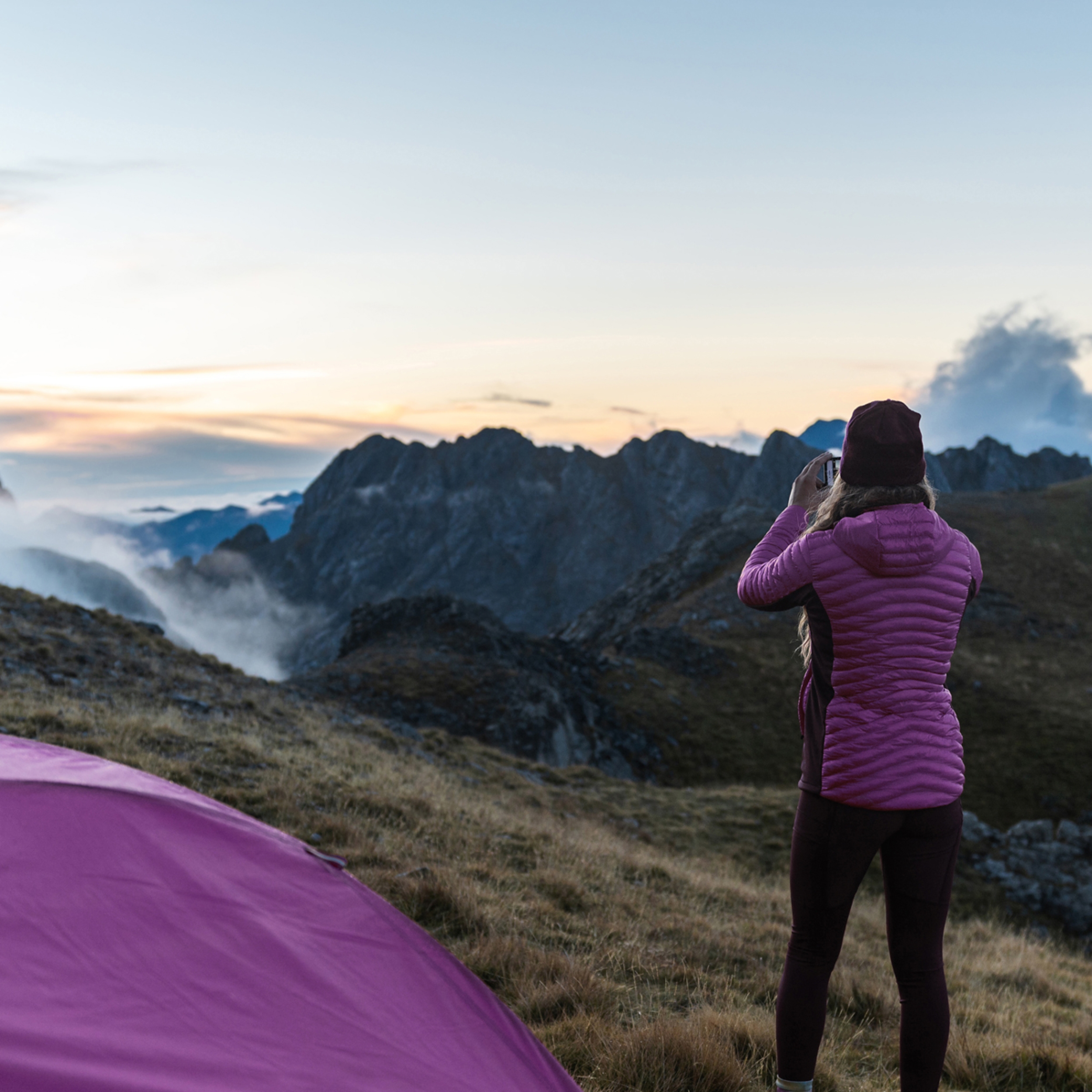 A woman is seen from the back wearing pink coloured hiking gear. She is taking a photo of the landscape with mountains and clouds in front of her. A pink coloured tent is seen in the corner of the photo.